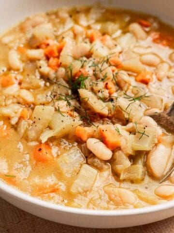A bowl of white bean and vegetable soup with carrots and onions, garnished with herbs, and a spoon resting inside.