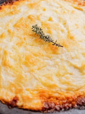 A baked shepherd's pie topped with golden mashed potatoes and garnished with a sprig of thyme on a round black dish.