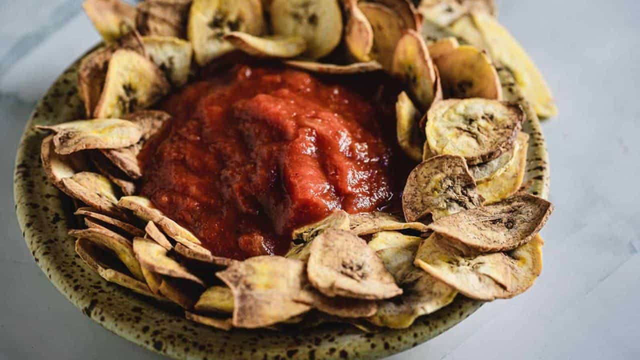 A speckled bowl filled with tomato sauce in the center, surrounded by a ring of crispy, thinly sliced plantain chips on a light surface.