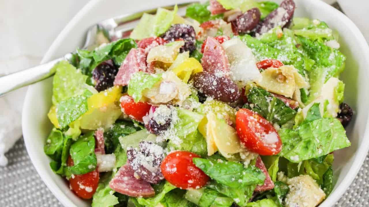 A bowl of salad with romaine lettuce, cherry tomatoes, salami, yellow bell peppers, black olives, grated cheese, and artichoke hearts. A fork is placed in the bowl.