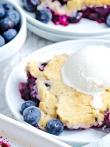 A plate with blueberry cobbler topped with a scoop of vanilla ice cream, fresh blueberries on the side, and utensils in the background.
