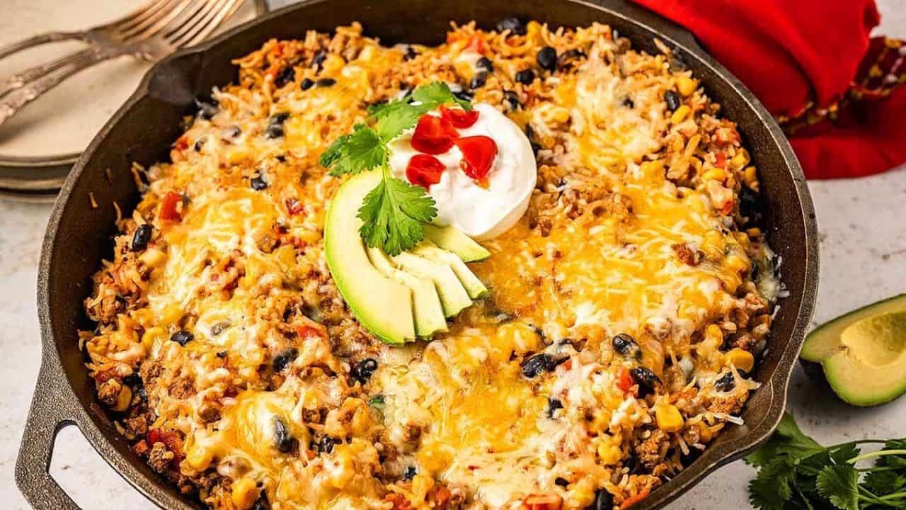 A skillet filled with a cheesy Mexican casserole topped with avocado slices, sour cream, cilantro, and chopped red pepper. Plates and a red napkin are in the background.