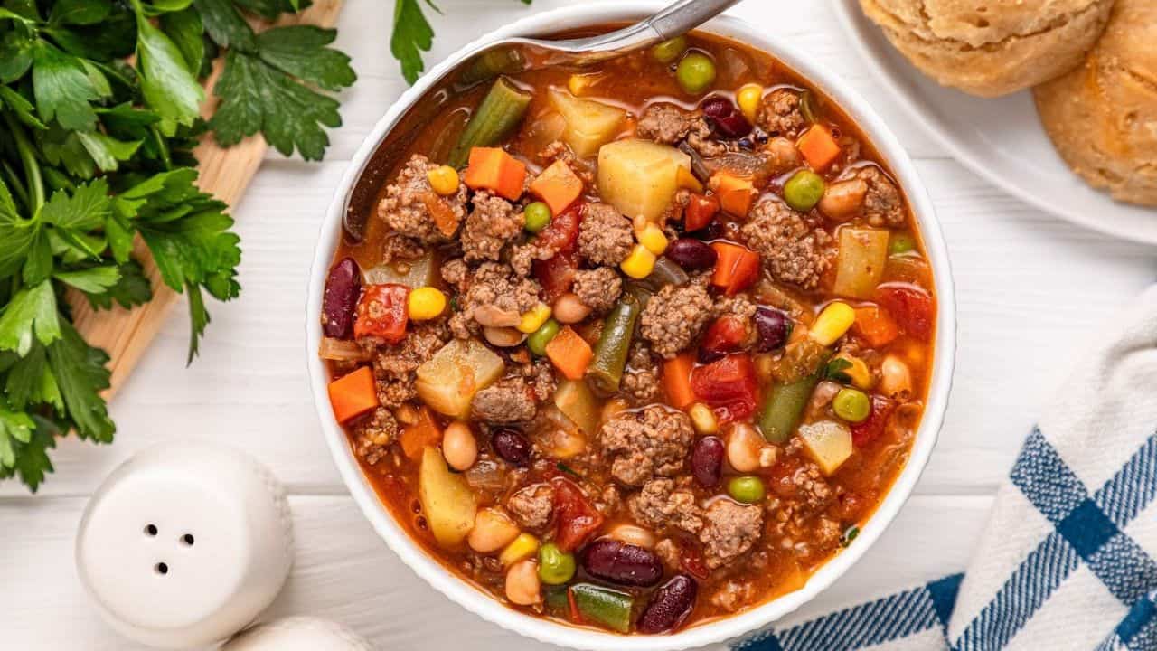 A bowl of hearty beef and vegetable soup with kidney beans, potatoes, carrots, peas, and corn, next to biscuits and parsley on a white table.
