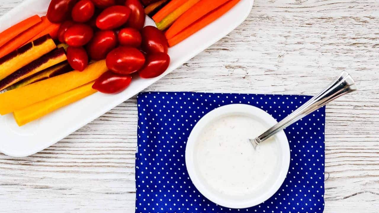A bowl of creamy garlic dressing with carrots and tomatoes on a blue cloth.