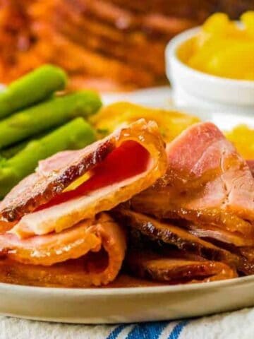 A plate with slices of glazed ham, steamed asparagus, and a serving of peaches in a small bowl, placed on a napkin.