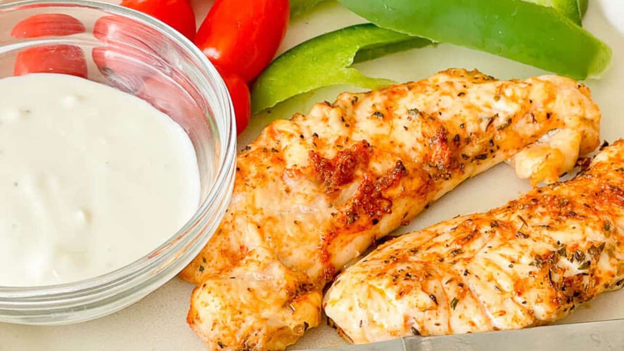Two seasoned, cooked chicken breast fillets on a plate with a bowl of white dipping sauce, sliced green bell pepper, and whole cherry tomatoes.