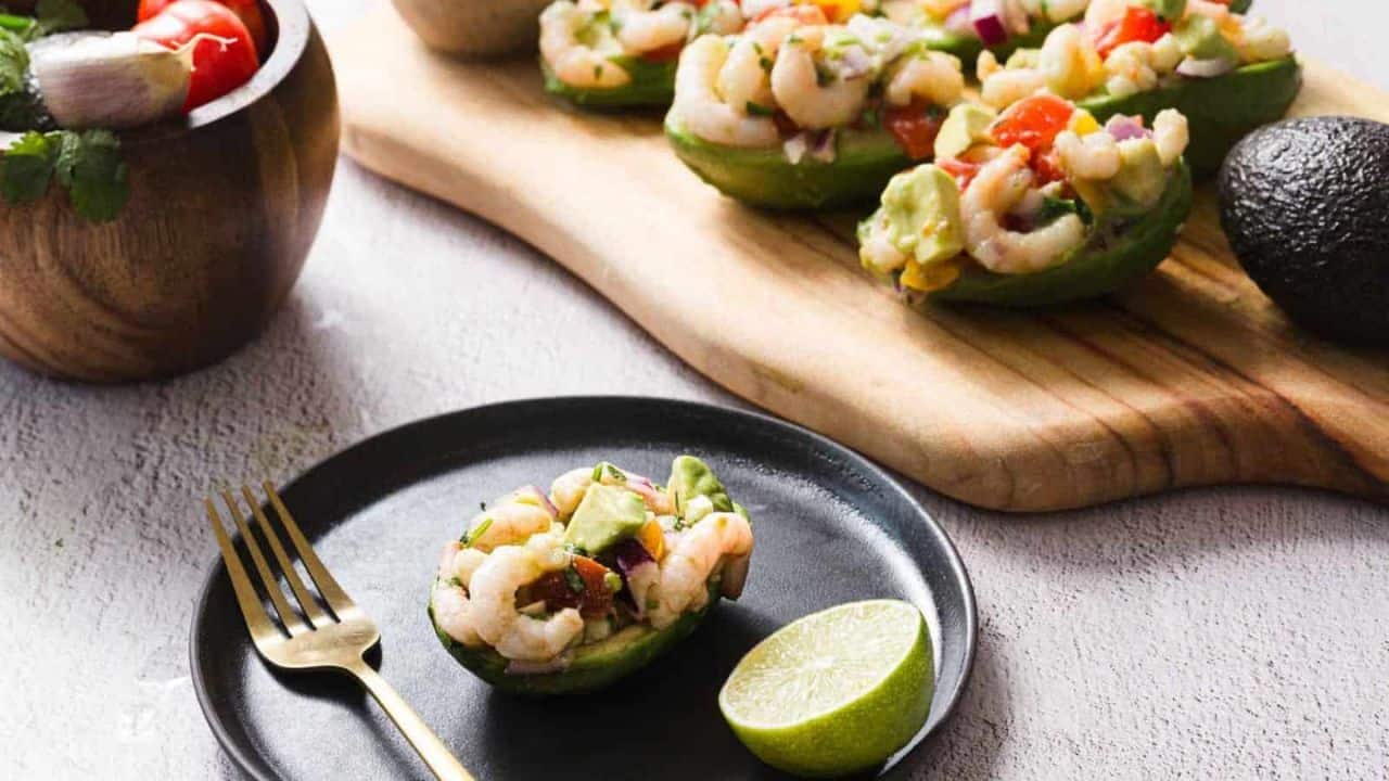 A black plate with a halved avocado filled with a shrimp salad, garnished with lime. A fork rests beside it. A wooden board holds more avocado halves in the background. A wooden bowl with mixed salad ingredients is partially visible.