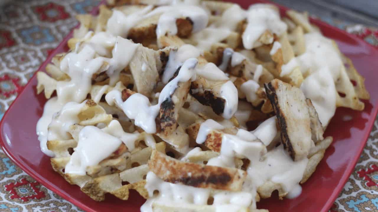 Waffle fries on a red plate topped with grilled chicken, alfredo sauce, and white cheese.