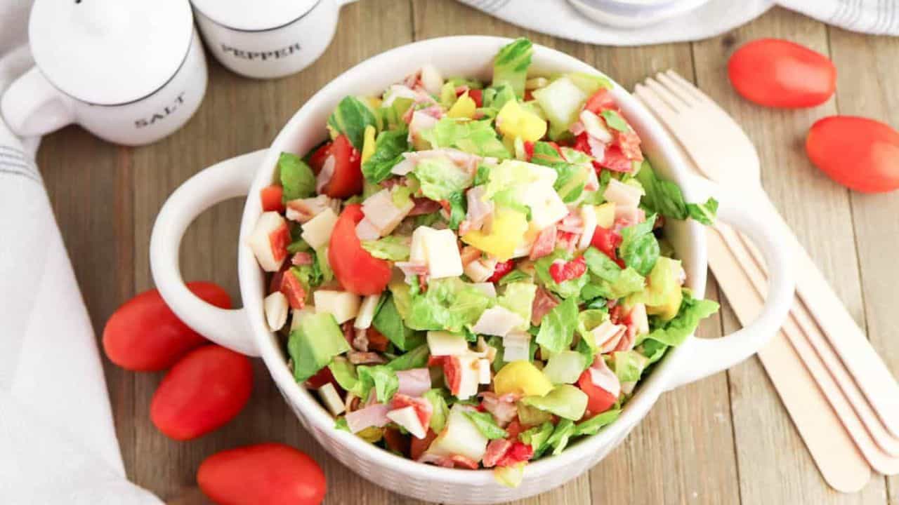A bowl of Grinder Chopped Salad with lettuce, tomatoes, cheese, and veggies sits on a wooden table, surrounded by grape tomatoes, a salt and pepper grinder set, and wooden utensils.