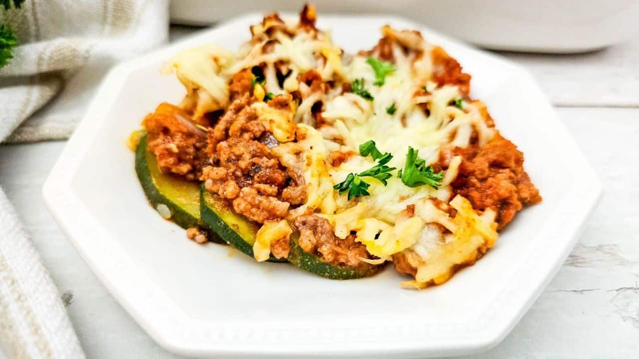 A serving of ground beef and zucchini casserole lasagna topped with melted cheese and parsley on a white plate.