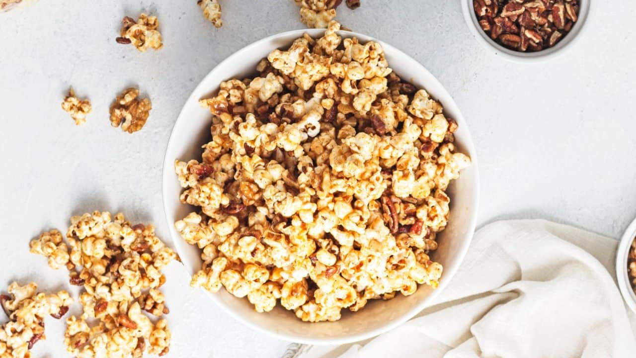 A white bowl filled with caramel popcorn and pecans sits on a light surface, with scattered popcorn and small bowls nearby.
