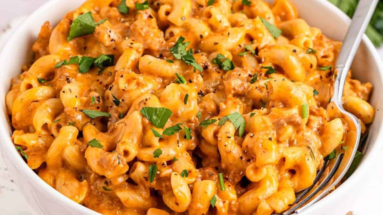 A bowl of creamy macaroni and cheese with ground meat, topped with chopped parsley and cracked black pepper, with a fork on the right side.