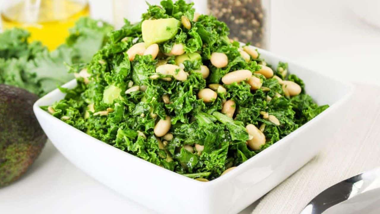 A square white bowl filled with kale salad, featuring beans and avocado pieces, placed on a table with a spoon beside it.