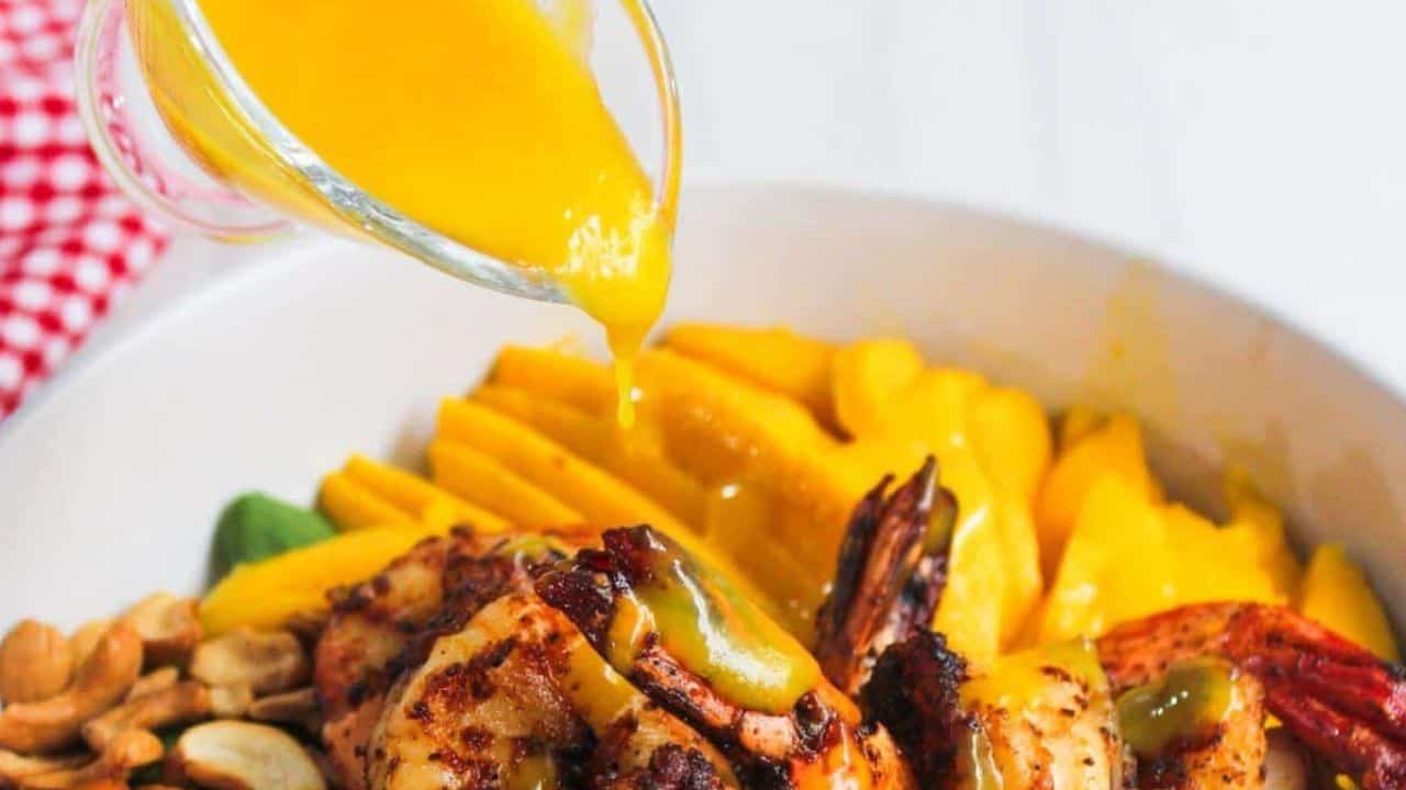 A glass container pours yellow sauce over grilled shrimp in a bowl with mango slices, nuts, and leafy greens.