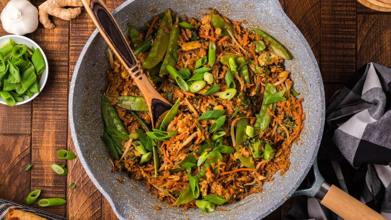 A pan filled with stir-fried rice, snow peas, shredded carrots, and chopped green onions, with a wooden spatula resting inside.