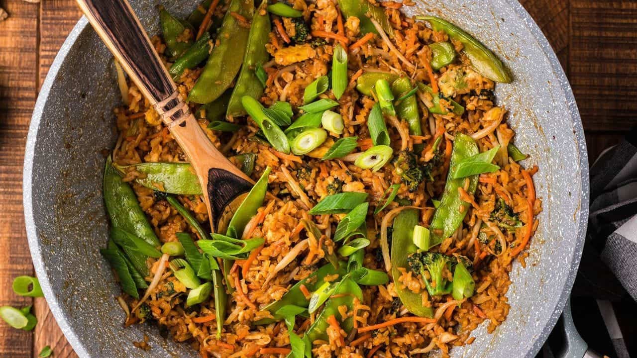 A skillet filled with fried rice, snow peas, shredded carrots, broccoli, and sliced green onions, with a wooden spoon resting inside.