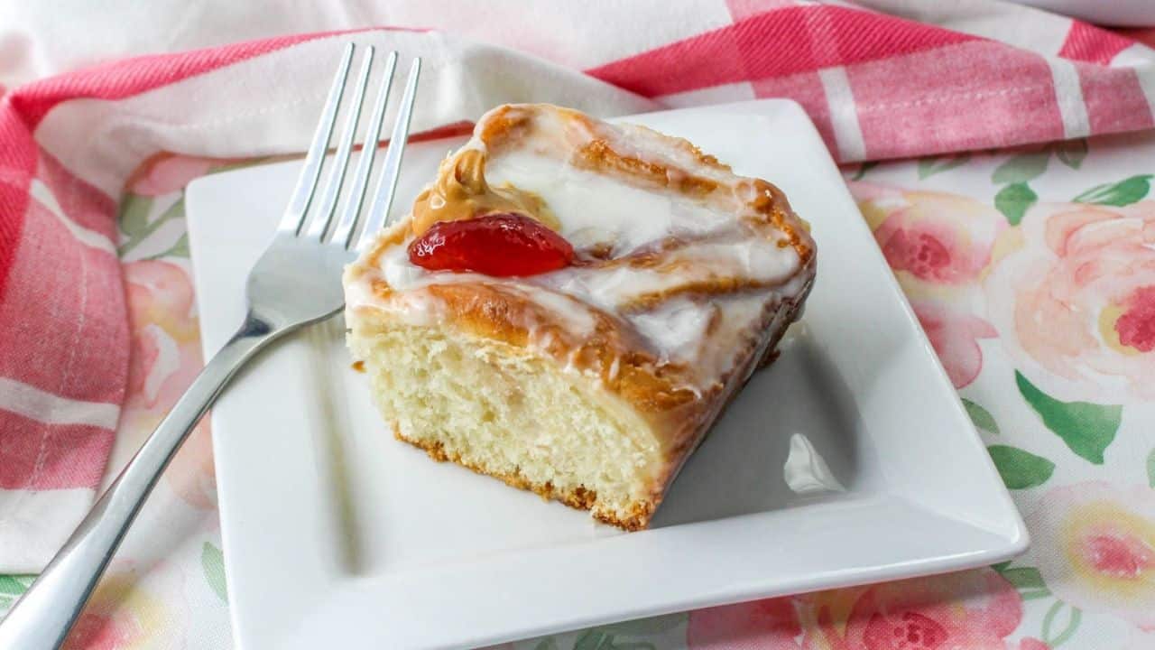 A slice of glazed peanut butter and jelly roll with a cherry on top sits on a white plate next to a fork, on a floral tablecloth.