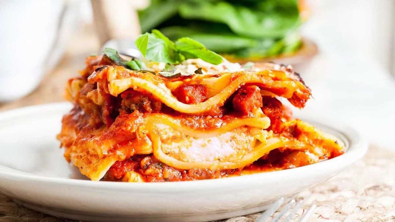 A slice of lasagna with layers of pasta, meat, tomato sauce, and cheese is served on a white plate, garnished with fresh basil.