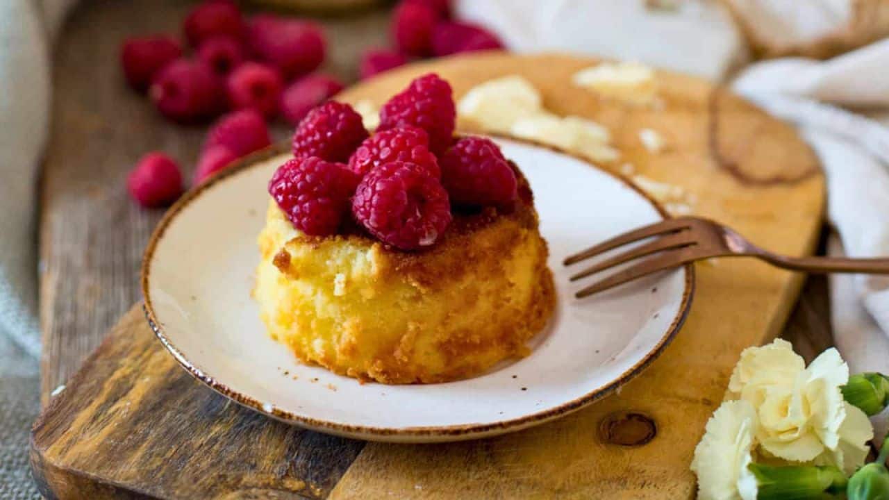 A fork holds a piece of cake above a white plate with a partially eaten slice of cake topped with raspberries. More raspberries and crumbs are scattered on a wooden board in the background.