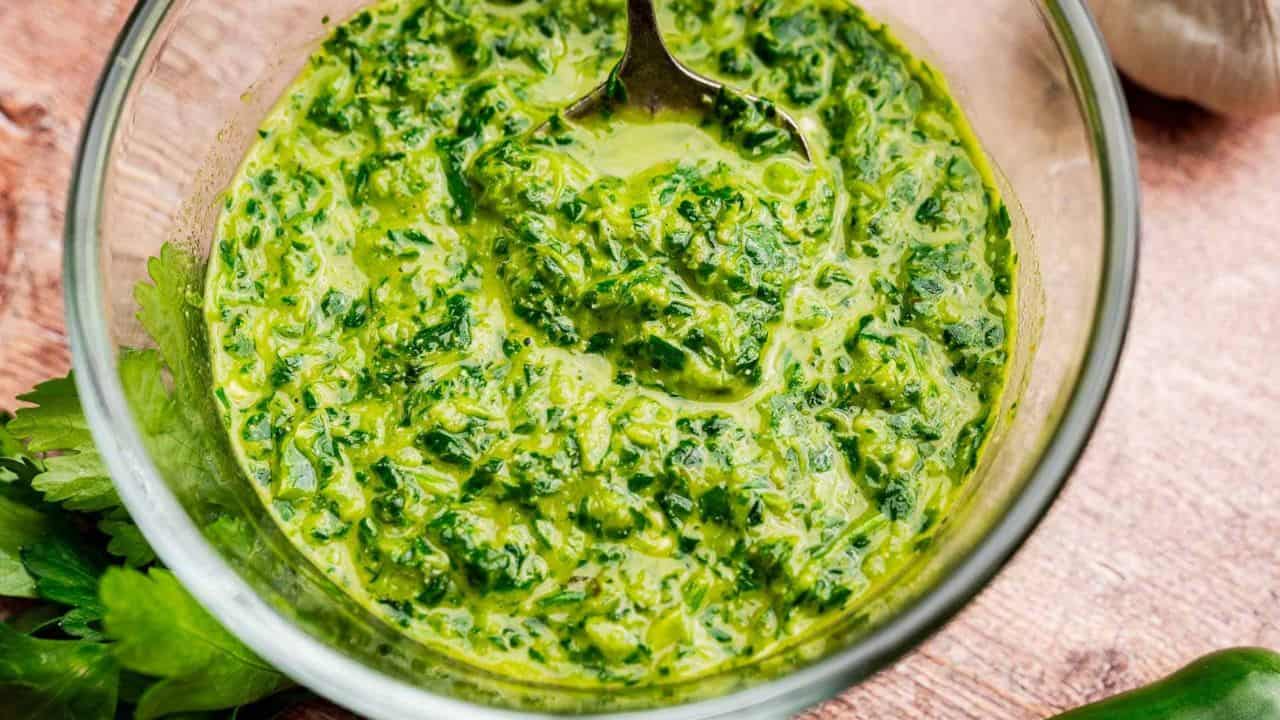 A glass bowl filled with green chimichurri sauce and a spoon, placed on a wooden surface with fresh herbs nearby.