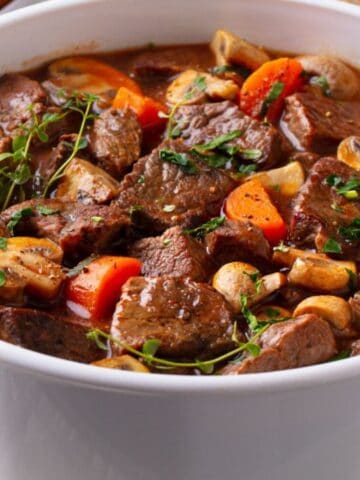 A white casserole dish filled with beef stew, featuring chunks of beef, mushrooms, carrots, and fresh herbs in a brown broth.