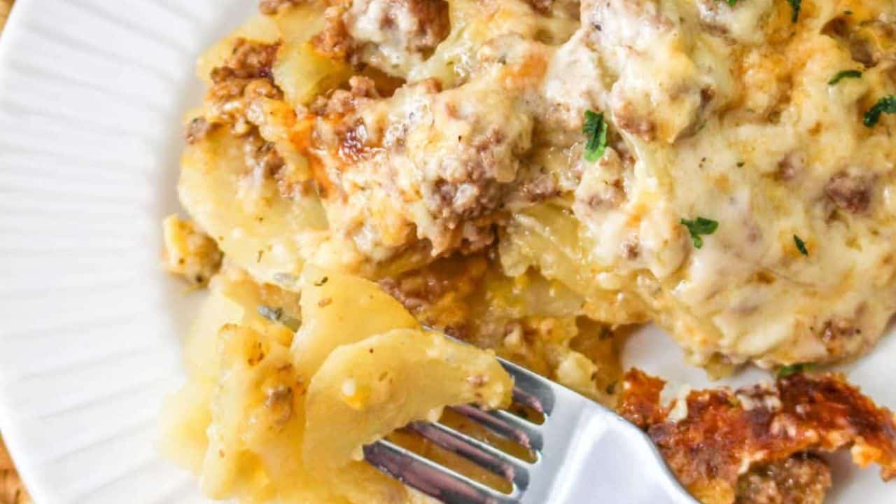 A plate of cheesy beef and potato casserole garnished with herbs, served with a fork. A potato, greenery, and a slow cooker are visible in the background.