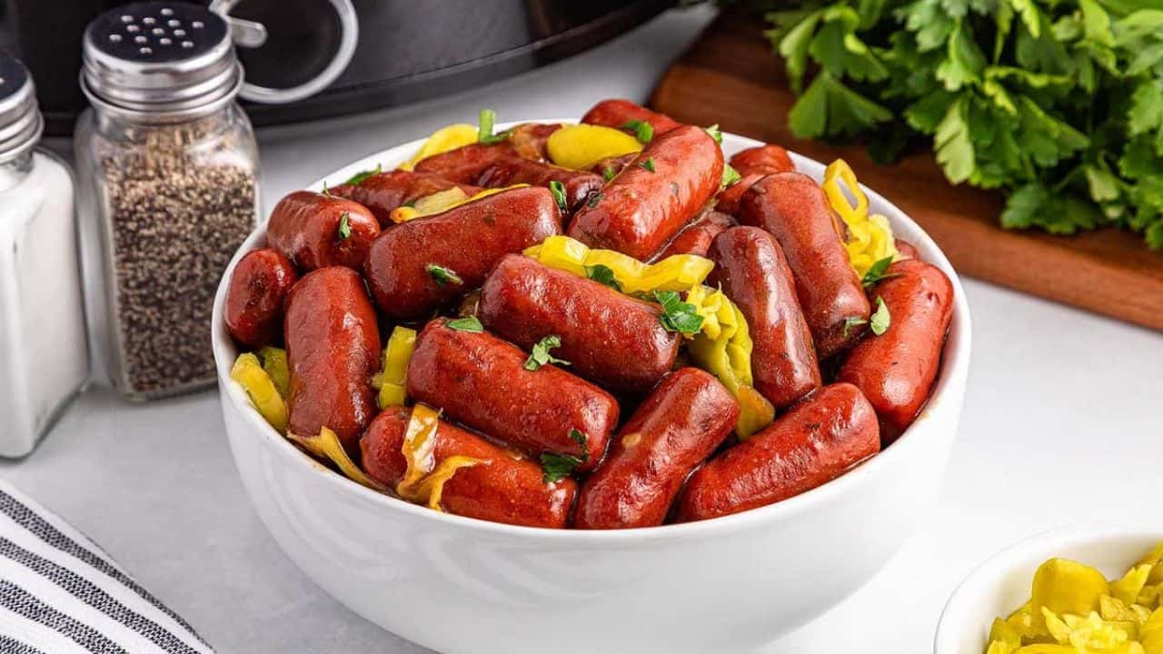 A white bowl filled with cocktail sausages garnished with chopped herbs and sliced peppers, placed on a countertop near salt and pepper shakers and fresh parsley.
