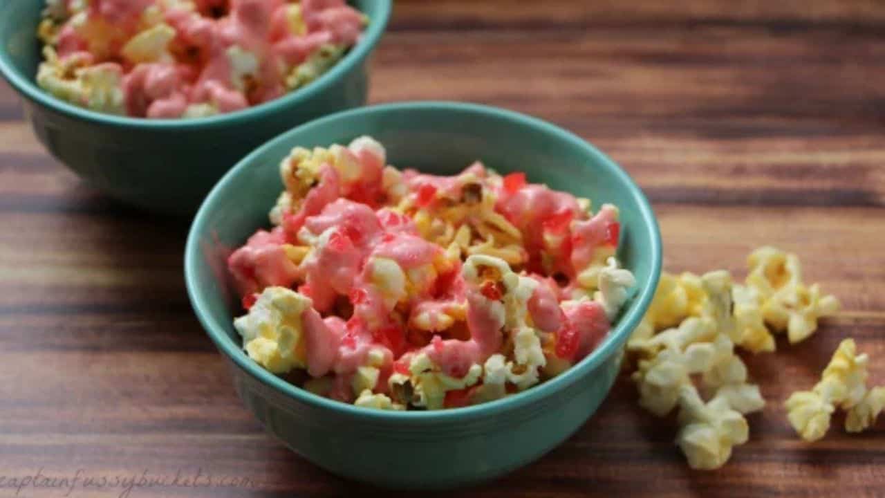Two teal bowls filled with popcorn drizzled with pink coating sit on a wooden surface, with a few pieces of plain popcorn scattered beside them.