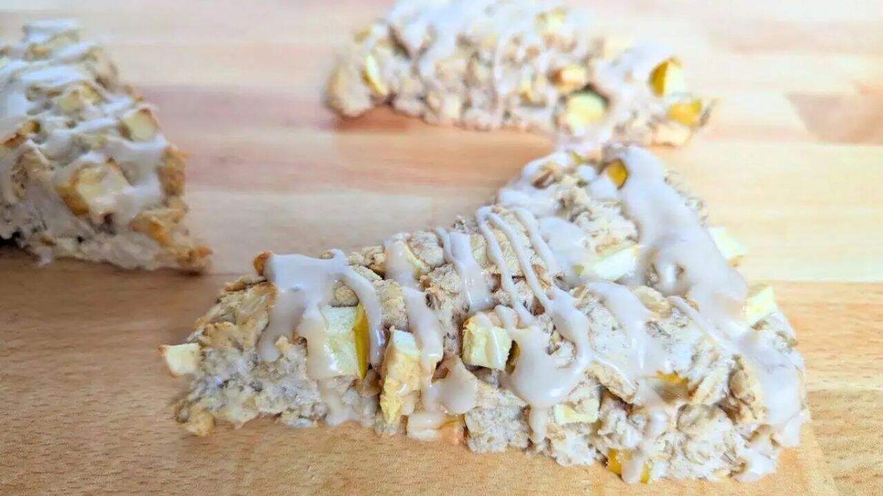 A triangular apple scone with visible oat and fruit pieces, topped with a light drizzle of white icing, on a wooden surface.
