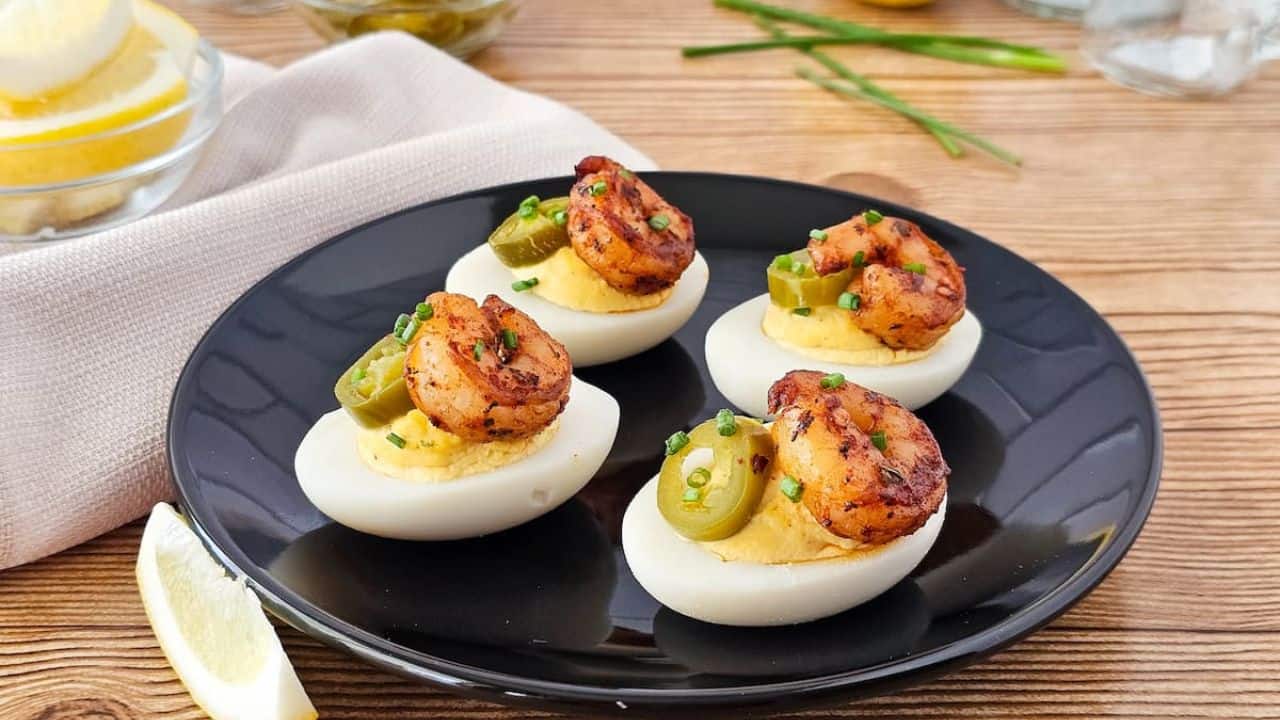 Cajun deviled eggs with shrimp are topped with sliced jalapeños, artfully arranged on a black plate set against a rustic wooden table, accompanied by a refreshing lemon wedge.