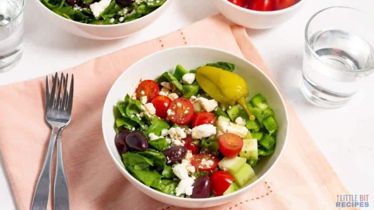A fresh greek salad with tomatoes, feta cheese, cucumbers, olives, and peppers, served with a fork and a glass of water on the side.