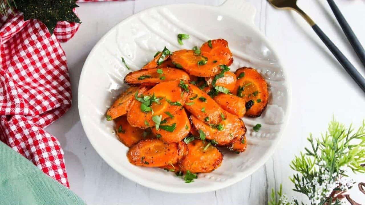 Sliced glazed carrots garnished with herbs on a fork, with more on a plate in the background.