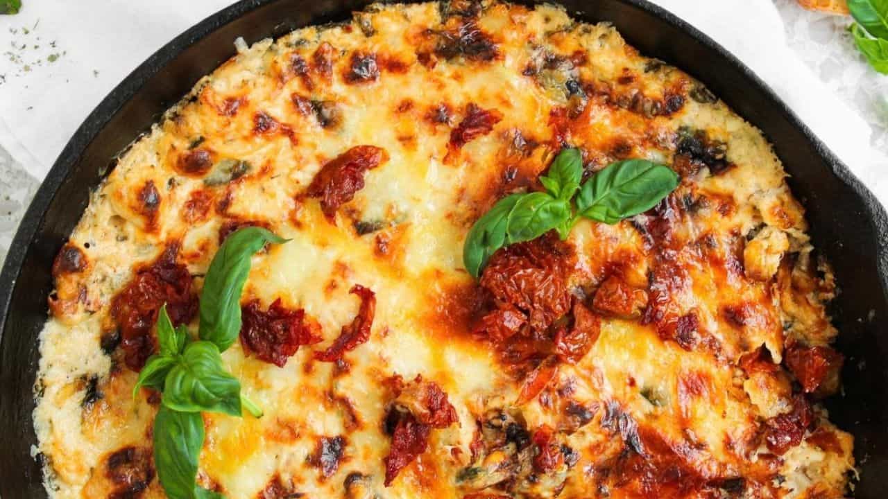 A baked cheese and tomato dish in a black skillet, topped with melted cheese, sun-dried tomatoes, and fresh basil leaves. Two slices of bread are placed beside the skillet on a white surface.