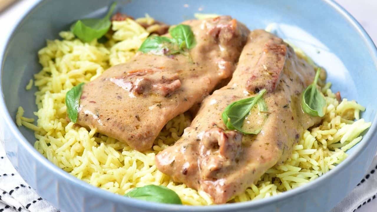 Two pieces of marry me chicken in a creamy sauce are served over yellow rice and garnished with fresh basil leaves in a blue bowl.