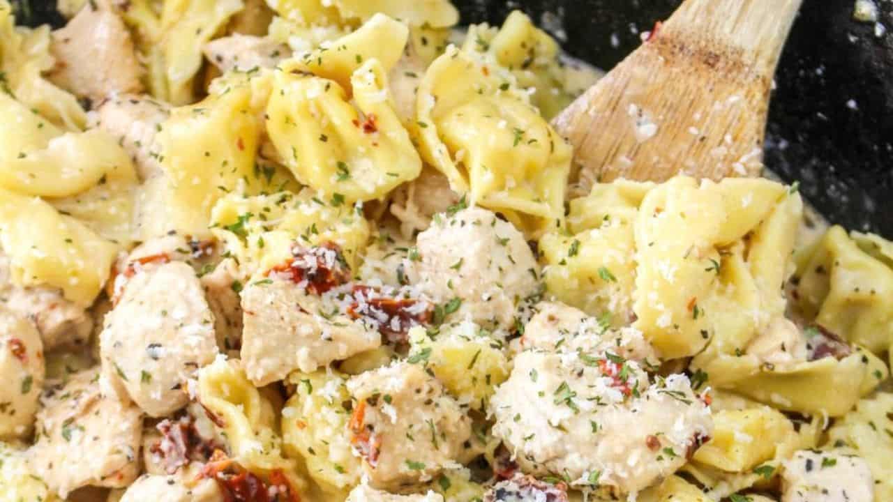 A white plate with creamy tortellini and chunks of seasoned chicken, garnished with herbs. A fork, napkin, and cheese grater are nearby on a striped cloth.