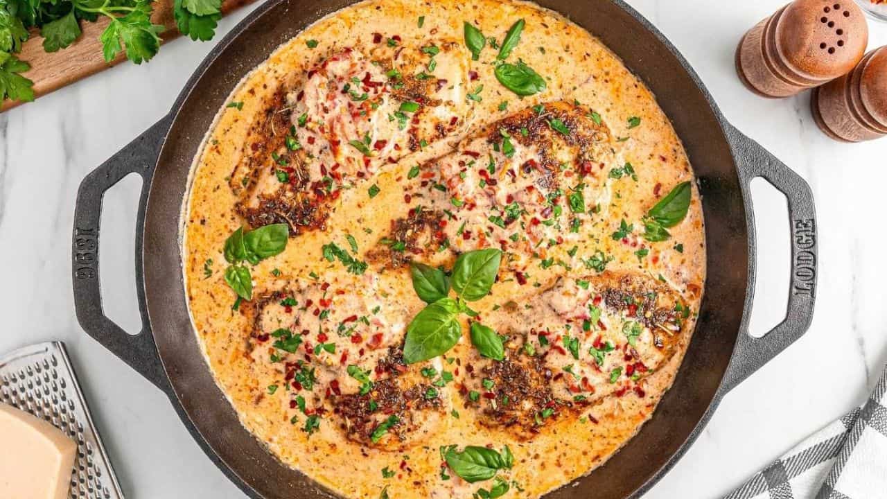 Four seasoned chicken breasts in a creamy Marry Me Chicken sauce, garnished with herbs and basil, cooked in a large cast iron skillet on a marble countertop.