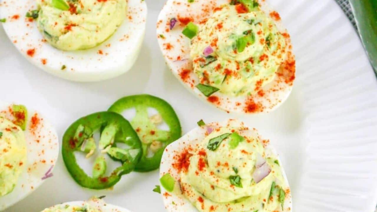 Plate of deviled eggs with avocado filling, garnished with paprika and green herbs, and sliced jalapeño.