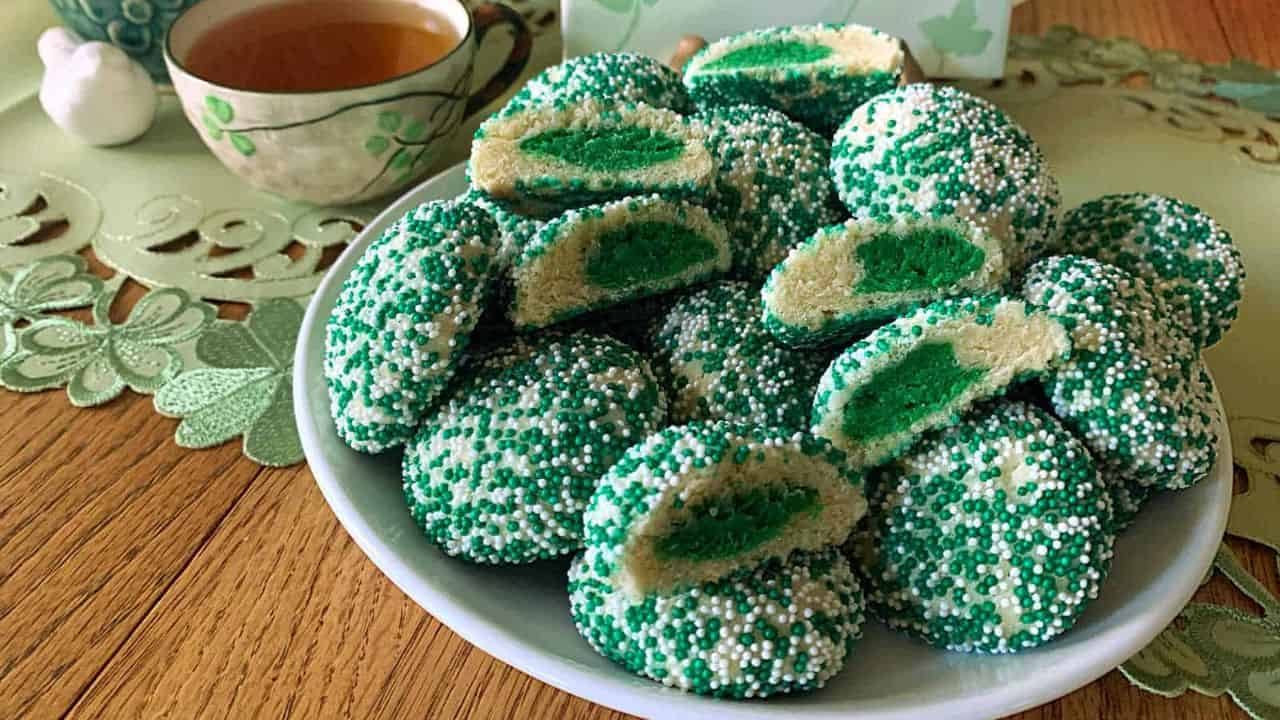 A plate of green and white sprinkled cookies, some cut in half to show green centers, next to a cup of tea on a table with a lacy placemat.