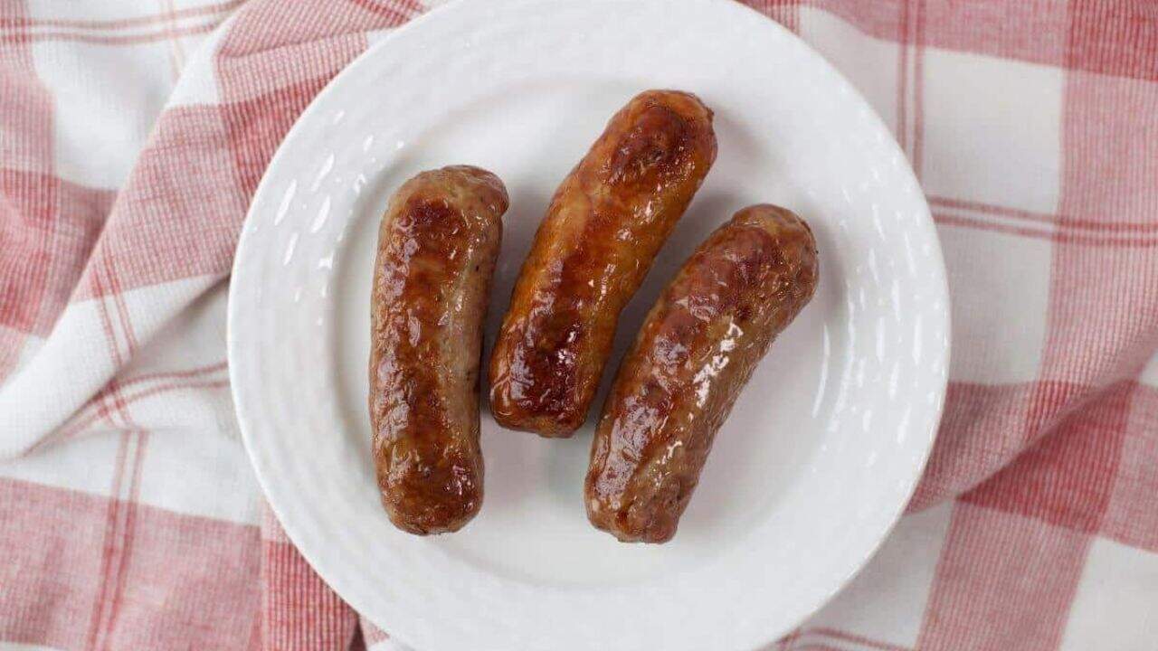 Three oven-baked sausages on a white plate with a red and white checkered napkin in the background.