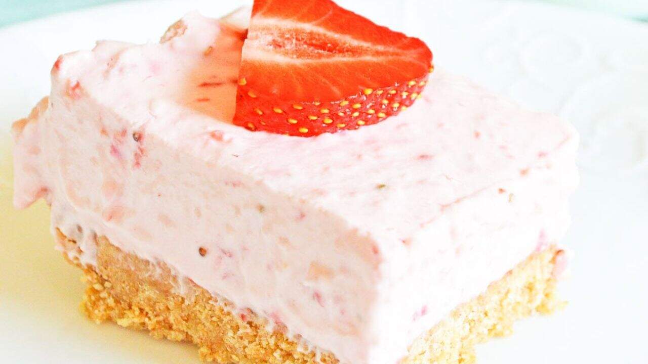A slice of strawberry cheesecake on a white plate, topped with a strawberry piece, with whole strawberries and a glass of milk in the background.