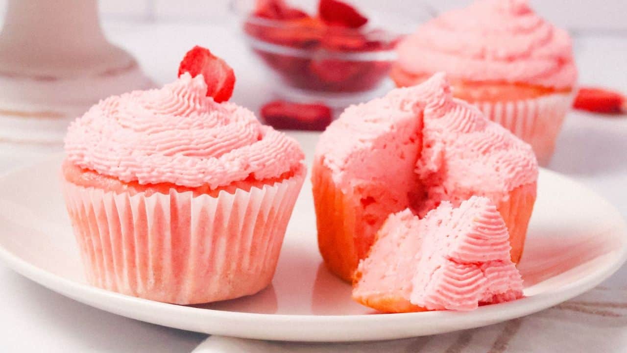 Two pink frosted strawberry cupcakes with luscious strawberry filling sit on a plate, one cut open to reveal the delicious inside. A blurred background subtly hints at more cupcakes and a bowl of strawberries, enhancing the sweet allure.