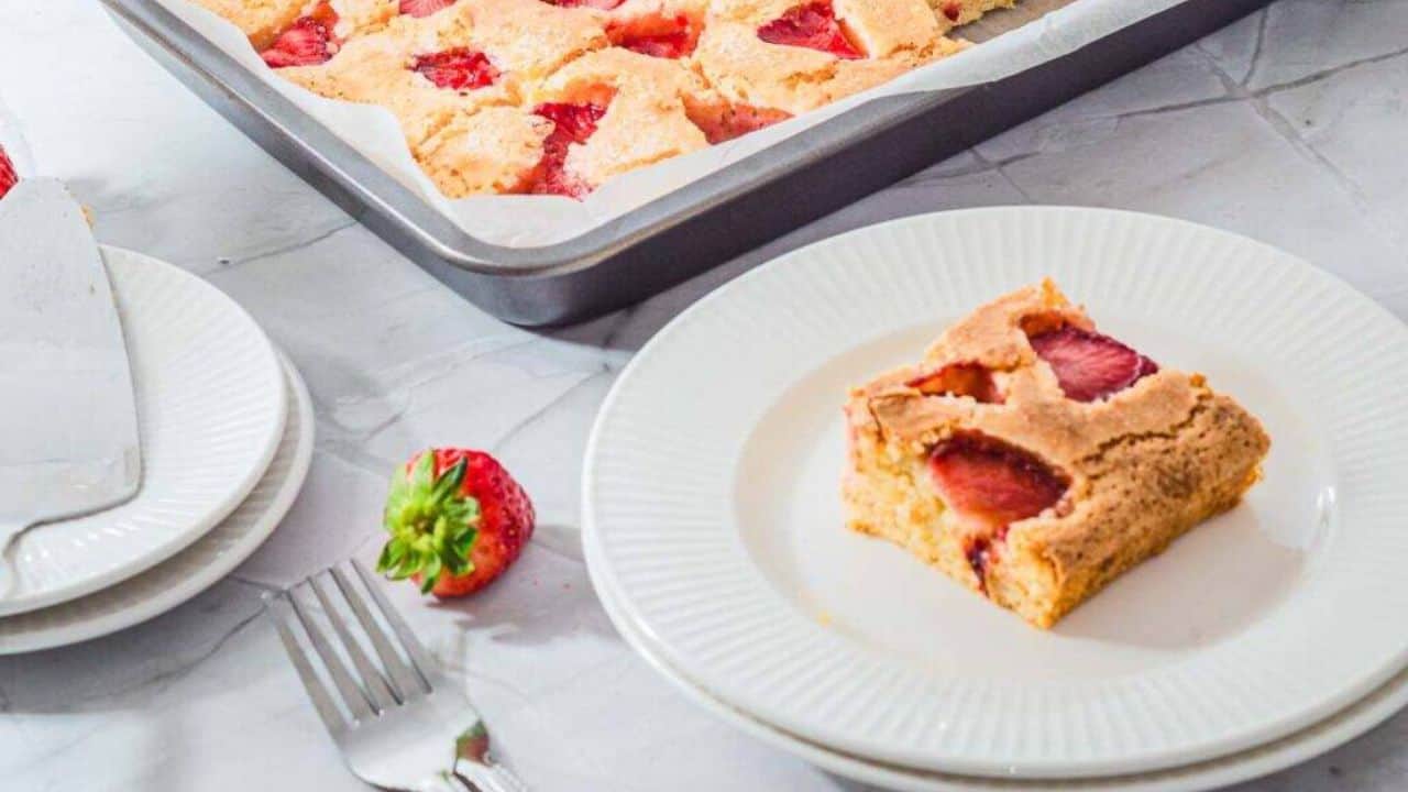 A slice of cake topped with strawberry sauce and a scoop of vanilla ice cream sits on a white plate, with a halved fresh strawberry beside it. A baking tray and whole strawberries are in the background.