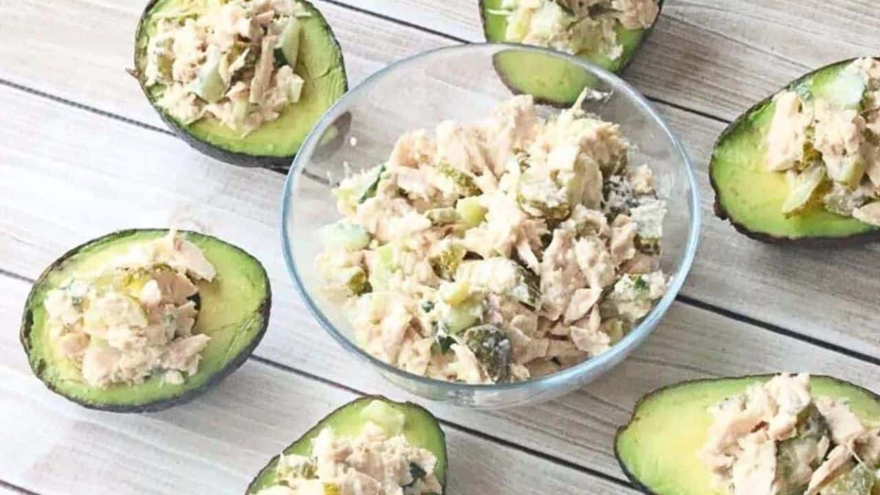 A glass bowl of tuna salad with pickles and multiple avocados stuffed with the salad around it.